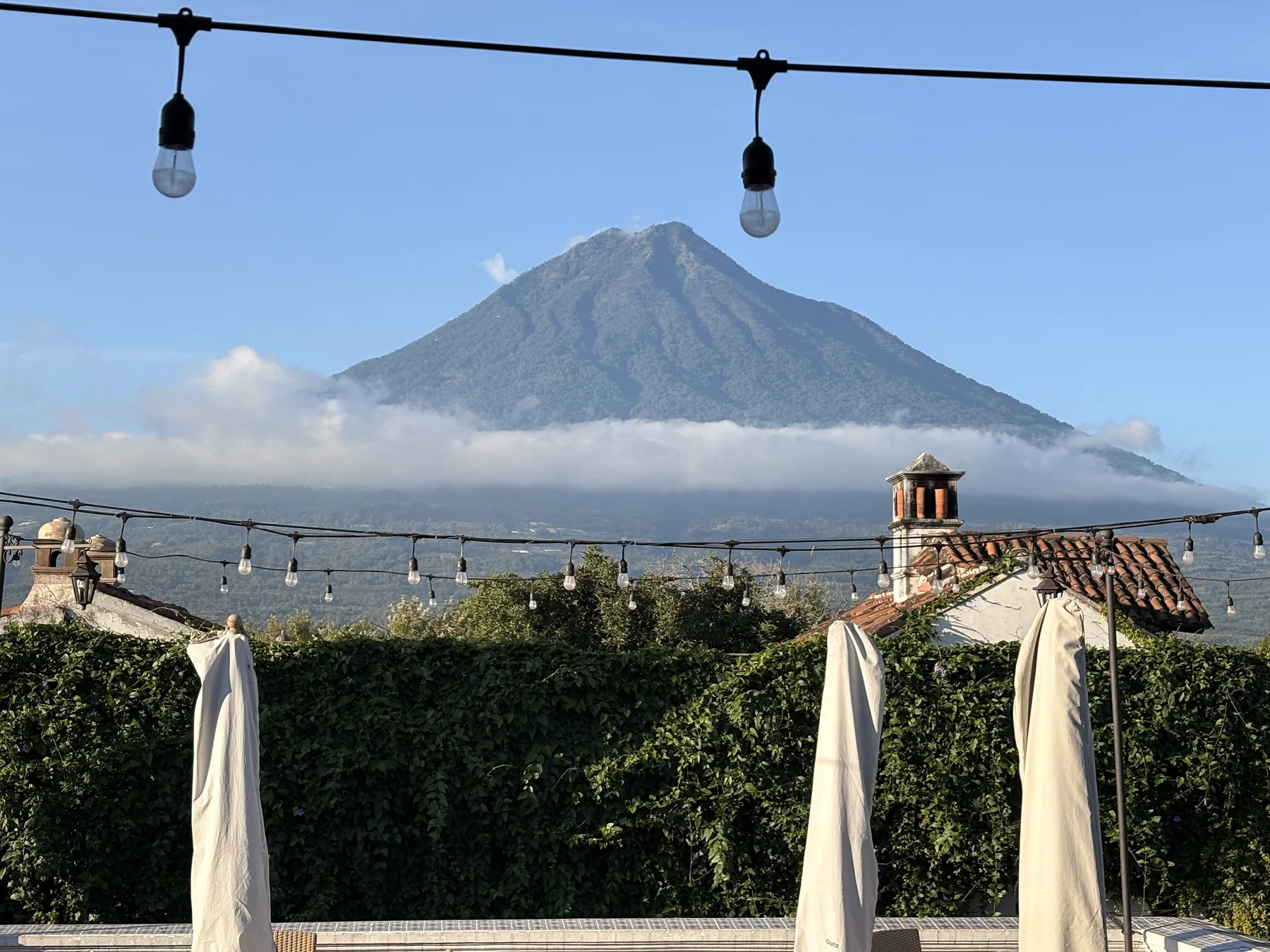 A volcano rising above a band of cloud, seen over tiled rooftops and a green hedge from a terrace in Antigua Guatemala
