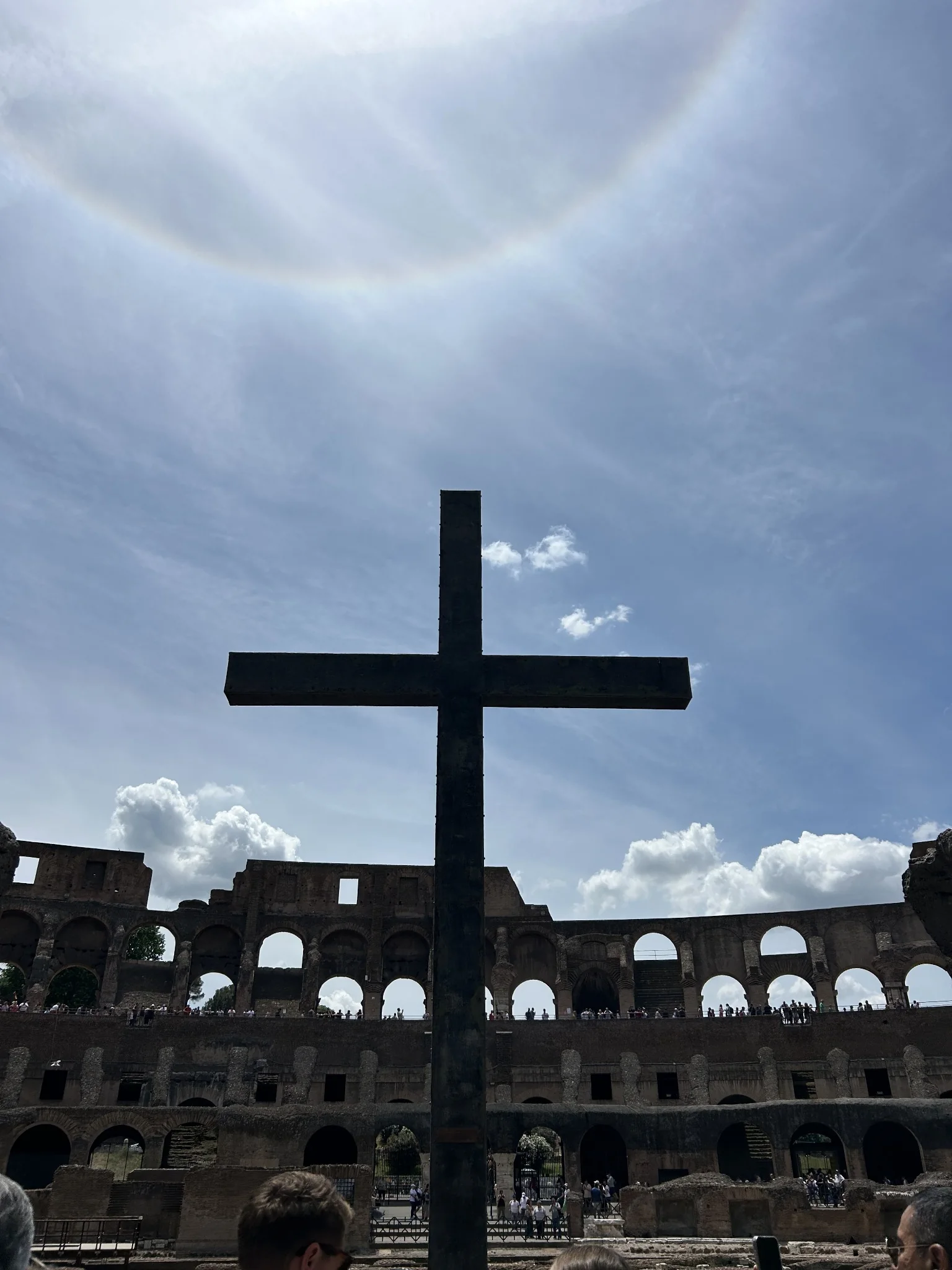 A tall dark cross inside the Colosseum in Rome, seen from below against the arches and a bright sky
